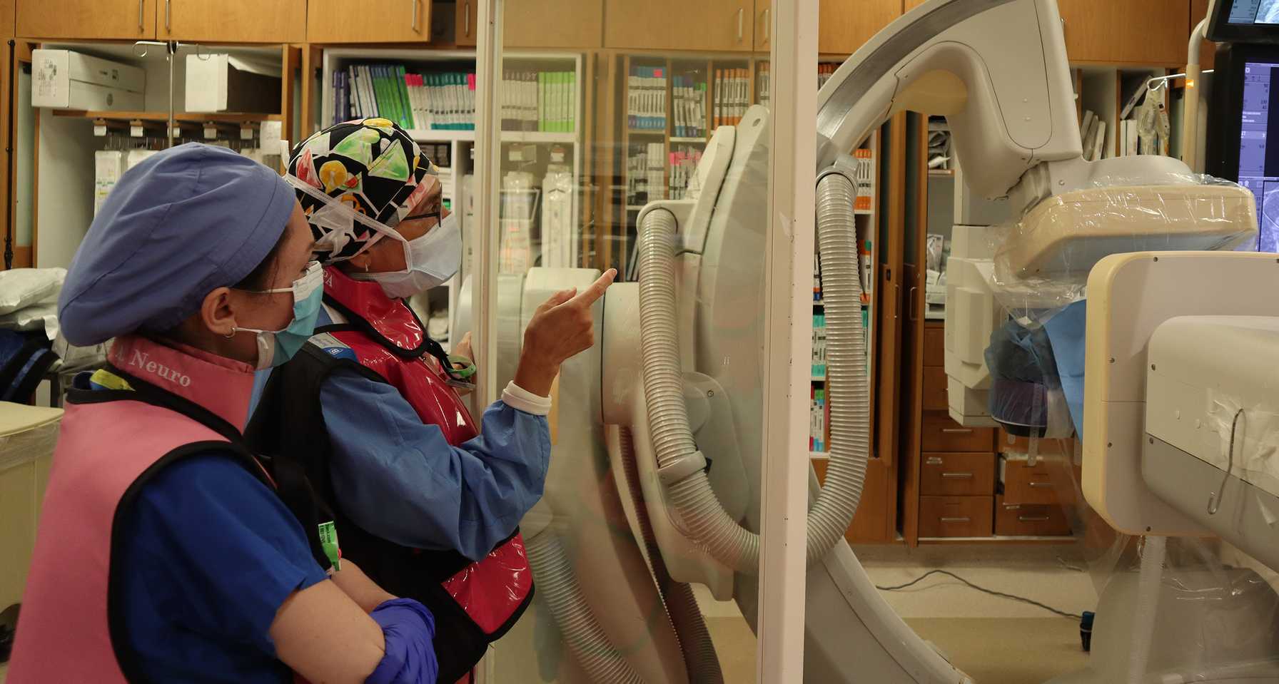 Two medical professionals in protective gear reviewing imaging scans on a monitor in a hospital procedure room, standing behind a clear protective shield next to advanced radiology equipment.