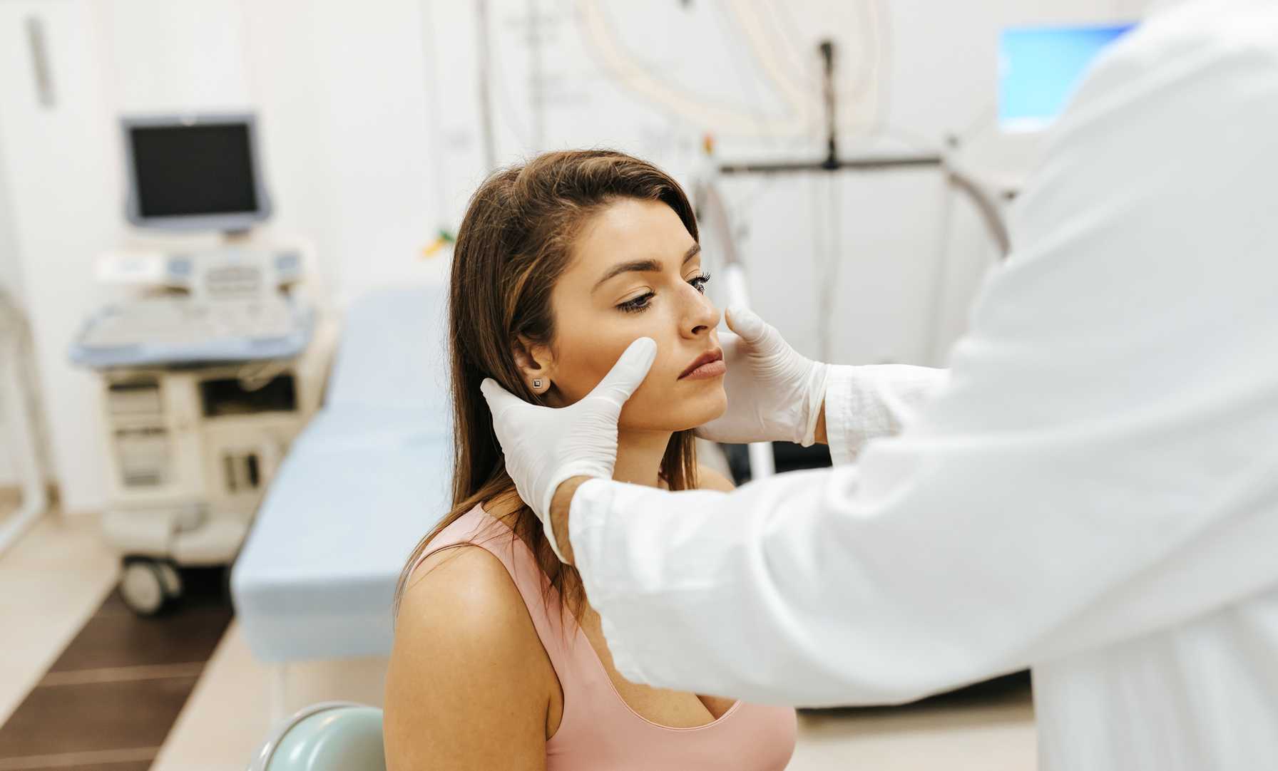 Doctor examining a woman's face in a medical office, with medical equipment and bed in the background.