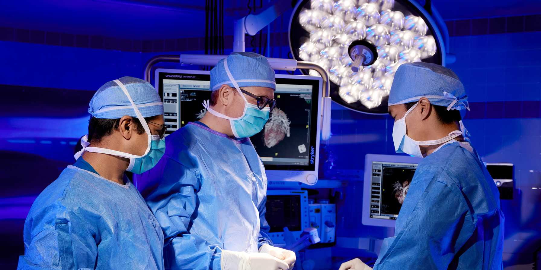Three surgeons in blue scrubs and masks working together in an operating room, surrounded by bright surgical lights and monitors displaying medical imaging.