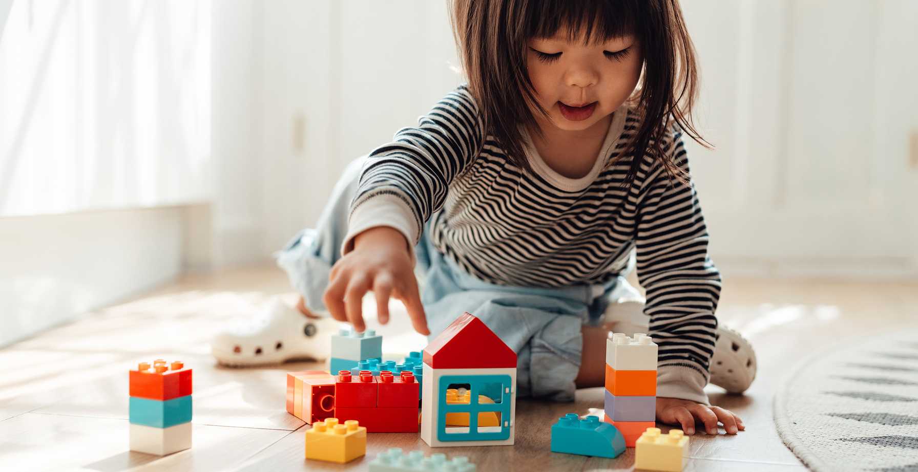 Young child playing on the floor with colorful plastic building blocks, wearing a striped shirt and light blue pants, in a sunlit room with wooden flooring.