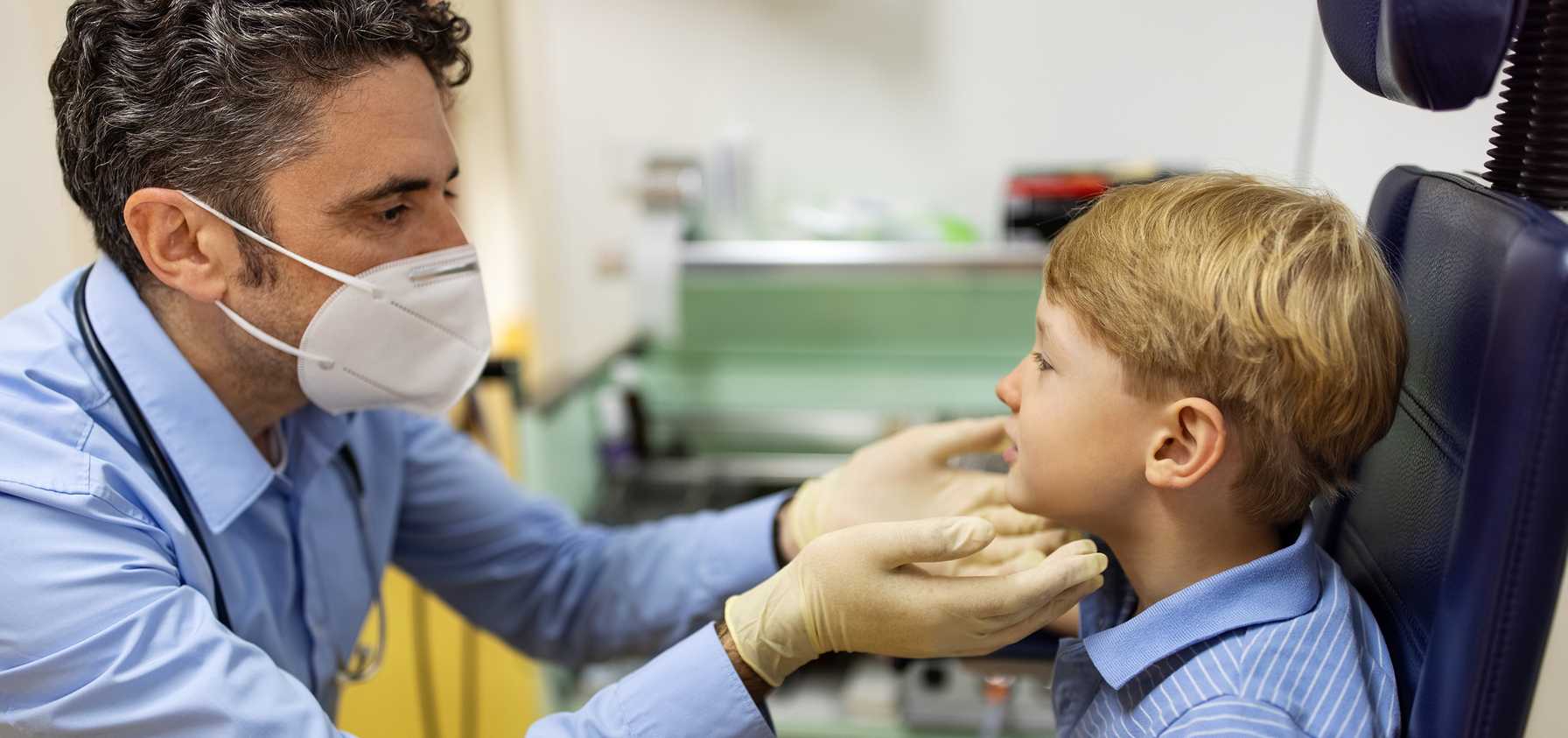 Male doctor wearing a face mask and gloves examining a young boy's throat in a medical office.
