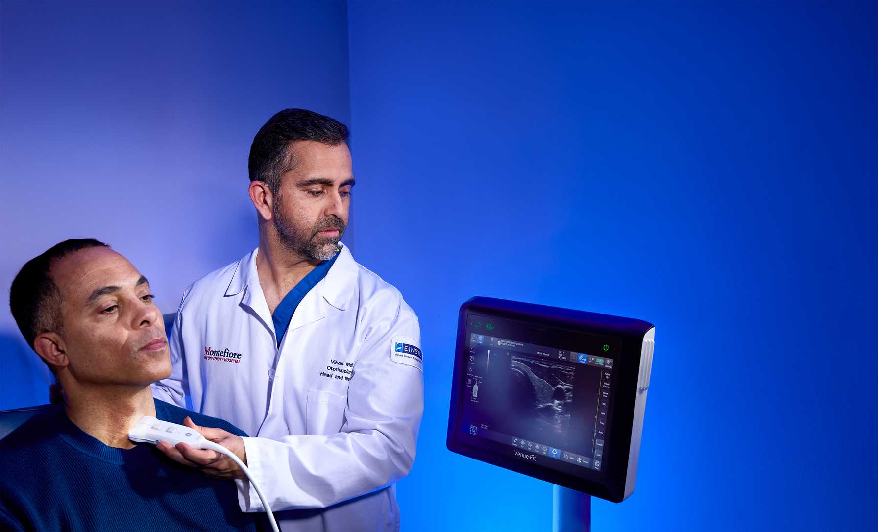 Doctor performing a neck ultrasound on a patient while viewing the results on a nearby monitor.